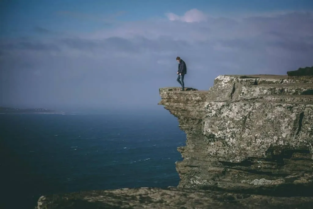 Courageous individual standing atop a rocky cliff with ocean view, embracing uncertainty, challenge, and freedom over the false security of a paycheck